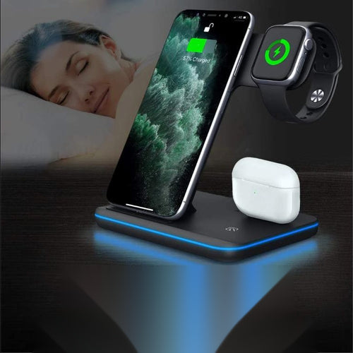 A black wireless charging stand with a blue LED light base, shown charging a smartphone and a smartwatch, with a white wireless earbud case resting on the stand. A blurred sleeping woman is visible in the background.